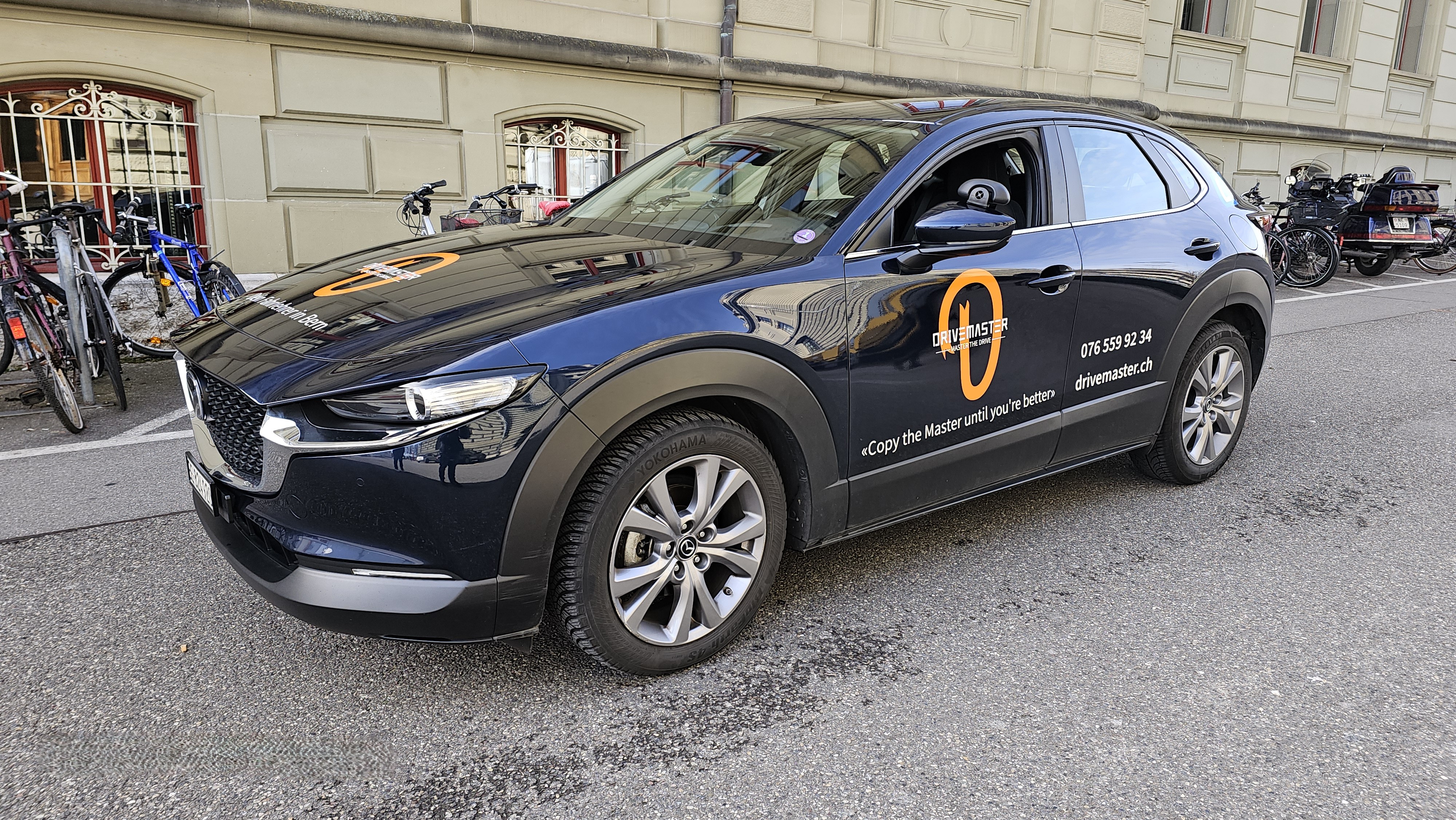 Drivemaster Fahrschulauto Mazda CX-30 AWD in Dunkelblau mit Firmenlogo, Telefonnummer und Slogan Copy the Master vor der Universität Bern – Fahrschule Bern